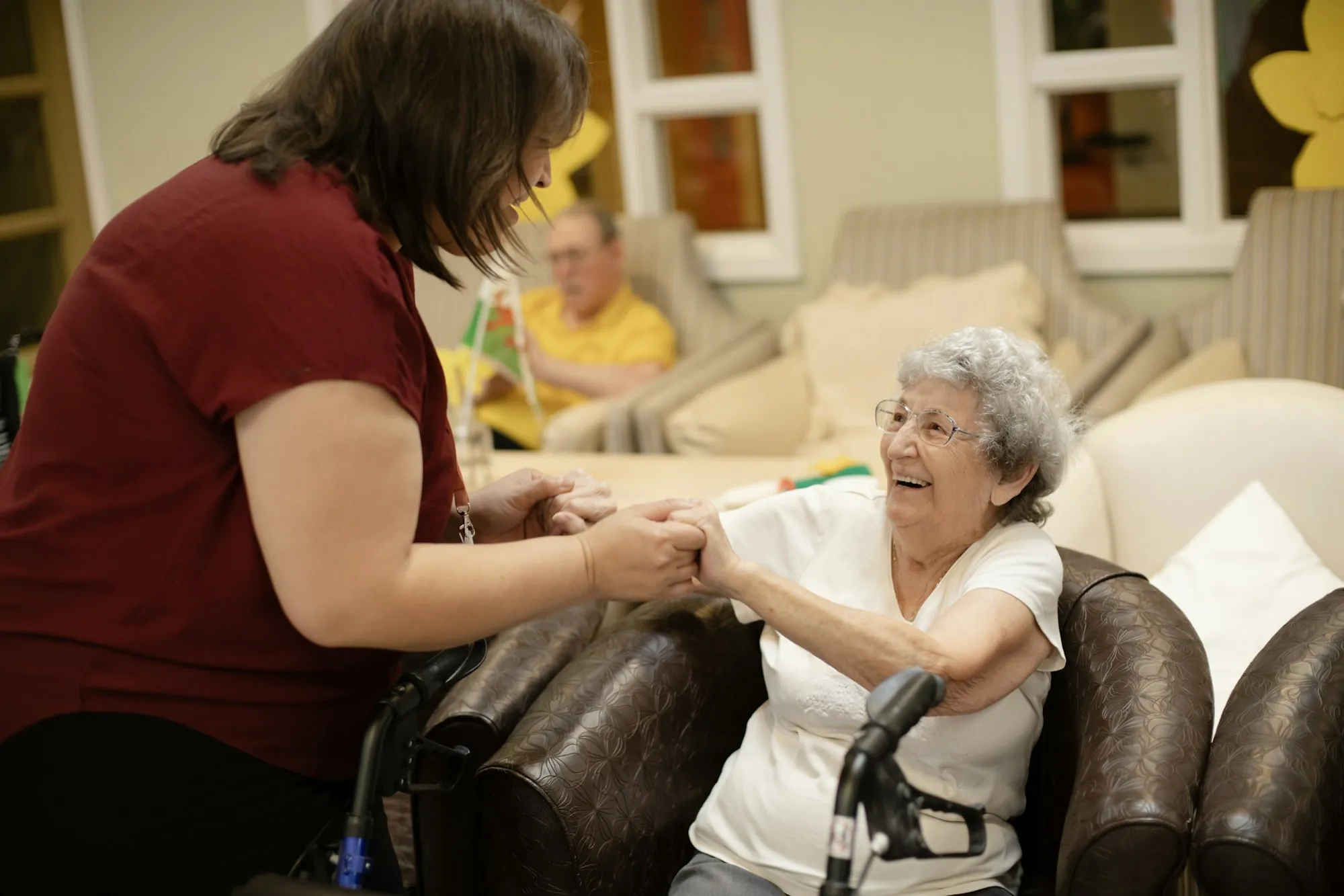 An aide supporting an older woman as she rises from a chair in a bright room