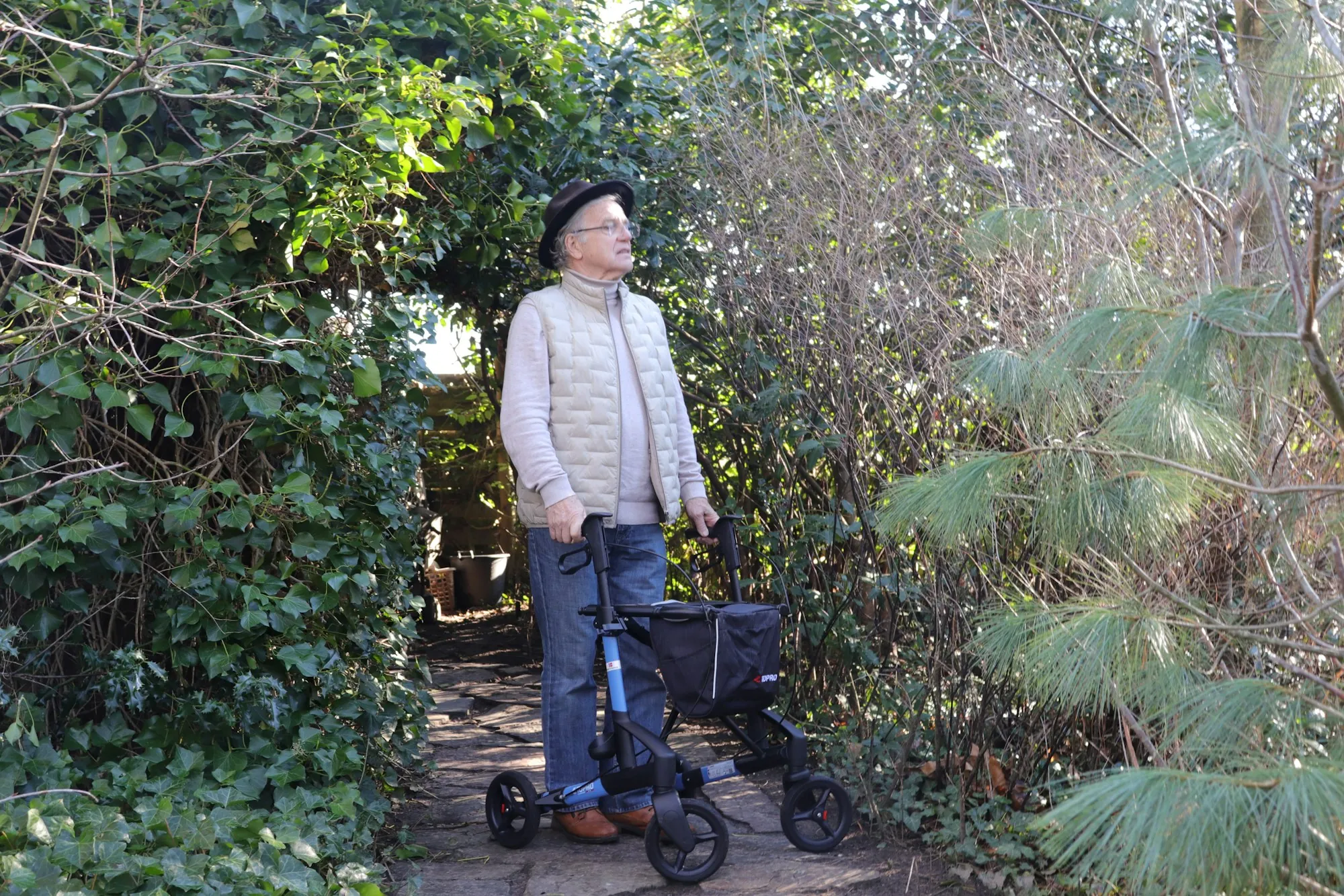 An older man walking a hallway with a wheeled frame while an aide walks beside him